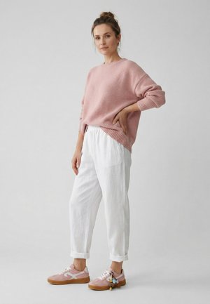 Frau steht mit der Hand in der Hüfte, trägt einen pinken Pullover, weiße Hose und pinke Sneaker mit einem Schlüsselanhänger vor einem schlichten grauen Hintergrund.