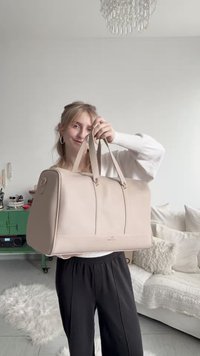 Ung kvinne innendørs som holder en stor beige skinnreiseveske, stående ved en koselig hvit sofa med teksturerte puter og et grønt skap i nærheten.