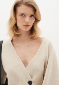 Cardigan de un bej deschis cu decolteu adânc în V, mâneci largi și un singur nasture negru. Material tricotat texturat, design minimalist.
