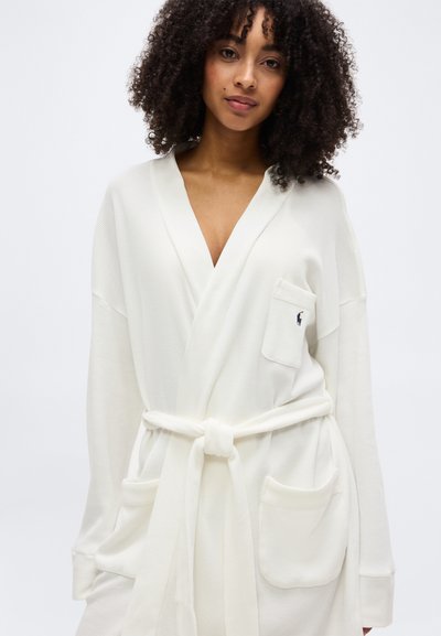 Γυναίκα με σγουρά μαλλιά που φοράει μια λευκή ρόμπα με ζώνη, μεγάλες μπροστινές τσέπες και ένα μικρό λογότυπο στην τσέπη του στήθους.