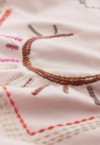 Zbliżenie na tkaninę z haftowanym wzorem słońca w brązowych, różowych i pomarańczowych nici na jasnym materiale.