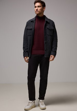 Donkergrijze wollen shirtjacket, kastanjebruine coltrui, zwarte broek en witte sneakers. Voorzien van dubbele borstzakken en een relaxte pasvorm.