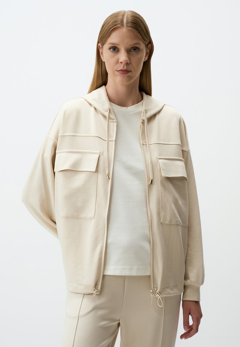 Beige gewatteerde jas met twee voorzakken, trekkoord aan de onderkant en zachte stof, gedragen over een lichte blouse en bijpassende broek.