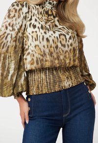 Blouse à imprimé léopard avec col montant, tissu transparent et taille froncée ; associée à un jean taille haute bleu foncé avec des accents métalliques.
