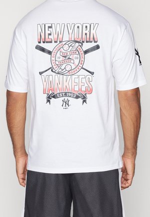 Homme portant un t-shirt blanc des New York Yankees avec des battes de baseball croisées et un logo de baseball dans le dos, associé à un short en mesh noir.