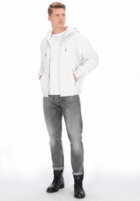 Ljusgrå zip-up hoodie i mjukt tyg, med framficka och dragsnören. Bärs över en vit T-shirt med grå jeans och svarta stövlar.