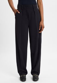 Wijdvallende pantalons van donkerblauw stof, met voorplooien, zijzakken en een platte tailleband. Vergezeld van zwarte instappers.