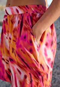 Žena s rukou v kapse voľných šortiek s výrazným ružovým, oranžovým a čiernym abstraktným vzorom, zobrazená od pása po stred stehien.