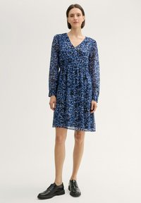 Blaues Blumenkleid aus transparentem Stoff, mit langen Ärmeln, einem V-Ausschnitt und einer gerafften Taille. Kombiniert mit schwarzen Schnürschuhen.