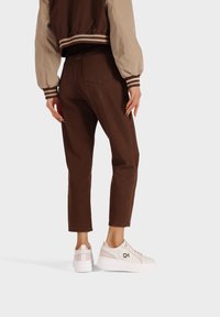 Pantaloni in denim marrone con vestibilità dritta, abbinati a sneaker bianche con un accento rosa. La giacca ha una manica di un marrone chiaro in contrasto.