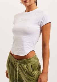 Valge sobiv T-särk lühikeste käiste ja ümmarguse kaelusega, millel on sädeleva detailiga "Juicy" logo. Kanduda oliiviroheline paelaga püksid.