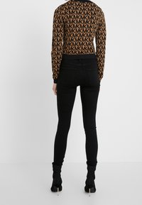 Jeans skinnies negros de tiro alto combinados con una blusa corta de manga larga con un patrón en tonos marrón y negro, que presenta un diseño de logo repetido.