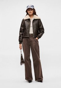 Casaco bomber de couro castanho com gola de pelo bege, combinado com calças castanhas de perna larga e uma blusa de cor clara. Inclui boné azul e mala.