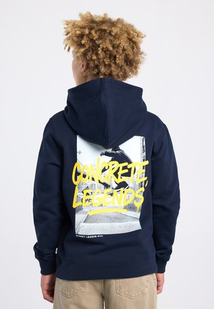 Personne aux cheveux bouclés portant un sweat à capuche bleu marine affichant une image de skateur avec le texte jaune "CONCRETE LEGENDS" au dos.