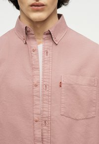 Άνδρας που φορά ανοιχτό ροζ πουκάμισο με κουμπιά και γιακά με κουμπί, με ορατή τσέπη στο στήθος στην αριστερή πλευρά.