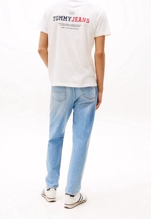 T-shirt en coton blanc avec l'inscription "TOMMY JEANS" en rouge et bleu marine au dos, associé à un jean en denim bleu clair et des baskets noir et blanc.