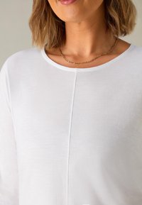 Top bianco a maniche lunghe realizzato in tessuto leggero, con scollatura rotonda e dettagli di cucitura discreti lungo la parte frontale. Collana a catena dorata visibile.