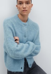 Donna con capelli corti che indossa orecchini a cerchio d'argento e un morbido cardigan blu chiaro con bottoni, braccia incrociate, su uno sfondo chiaro e semplice.