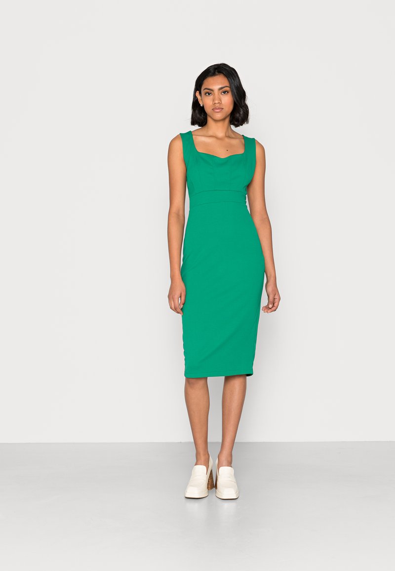 Vestido midi verde, ajustado, com decote quadrado, design sem mangas, textura suave e silhueta até ao joelho, combinado com sapatos claros.