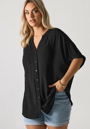 Blonde Frau trägt eine lockere schwarze Bluse mit Knöpfen und hochgekrempelten Ärmeln sowie hellblaue Jeansshorts, posiert mit einer Hand in der Tasche.