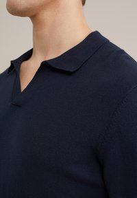 Suéter polo de punto azul marino con una textura suave, que presenta un cuello clásico y un diseño de escote en V. Costuras limpias y detalles mínimos.