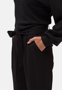 Svart topp med prickig textur som har en fluga i midjan ovanpå svarta byxor med fickor, som visar upp ett smidigt material och en strömlinjeformad design.