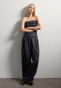 Una donna bionda si trova contro un muro bianco con le braccia incrociate, indossando un top nero senza spalline, pantaloni neri a gamba larga e scarpe nere con punta.