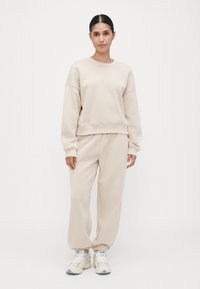 Kvinne som står iført beige oversized sweatshirt, matchende joggebukser og hvite joggesko mot en ensfarget hvit bakgrunn.