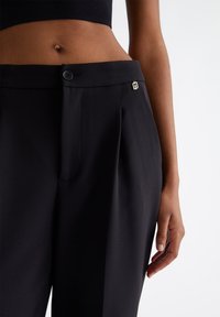 Pantaloni neri su misura con una texture liscia, dotati di chiusura a bottone e pieghe sottili. Include un piccolo dettaglio del logo metallico.