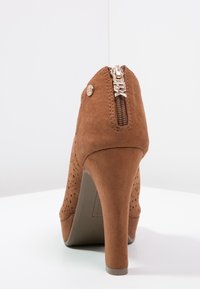 Zapato de tacón alto de ante marrón con detalles perforados y un zipper en la parte trasera. Presenta una punta redondeada y un tacón delgado y texturizado.