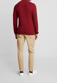 Pullover rosso a maglia con superficie tessurizzata, maniche lunghe e colletto alto, abbinato a chinos beige e sneakers bianche.
