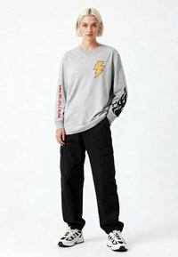 Junge Frau mit kurzen blonden Haaren, die einen grauen Pullover mit Blitz- und Flammenmotiven, schwarze Cargo-Hosen und weiße Sneaker trägt.