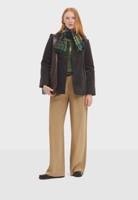 Schwarze Cordjacke, grüner Pullover, karierter Schal, beige weit geschnittene Hose und braune Slipper. Das Model hält eine braune Handtasche.