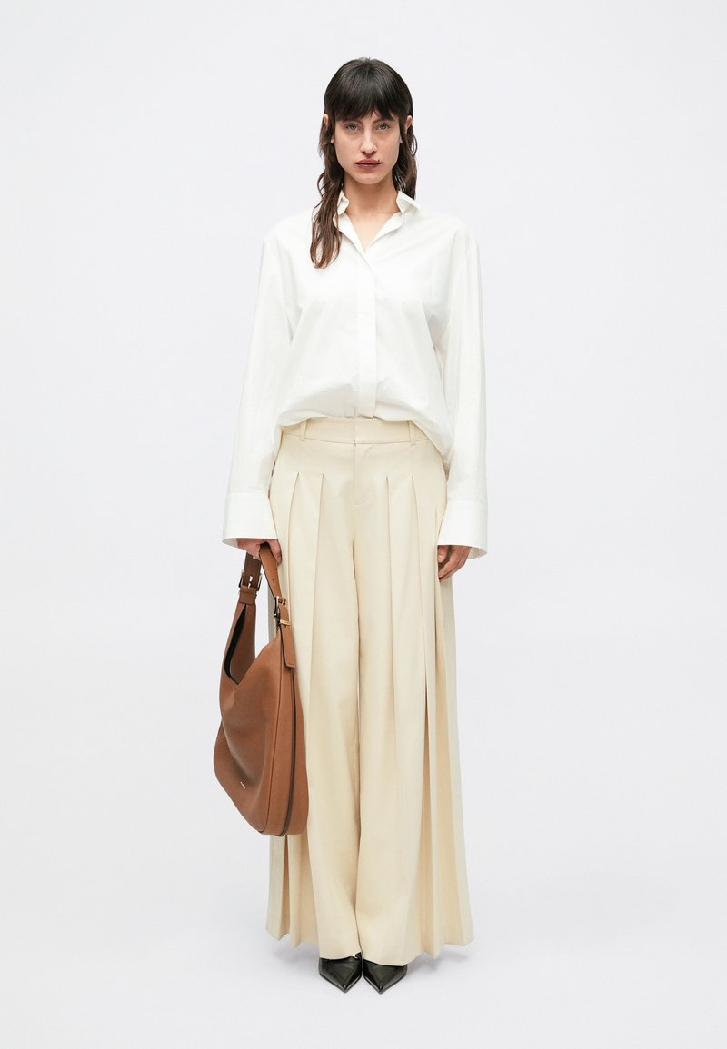 Camisa blanca abotonada, pantalones beige plisados, zapatos negros puntiagudos y bolso marrón de hombro. Diseño minimalista y paleta de colores neutros.