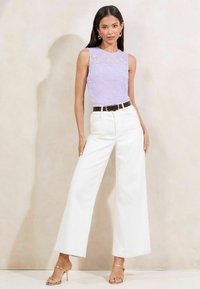Top senza maniche in pizzo lavanda con scollatura alta, abbinato a pantaloni bianchi a gamba larga e una cintura nera. Sandali con tacco dorati completano il look.