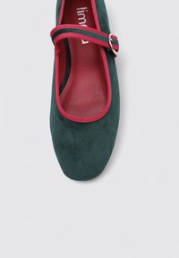 Zapato plano verde de ante con puntera redondeada y una correa rosa en contraste que cruza el pie, con una pequeña hebilla de plata.