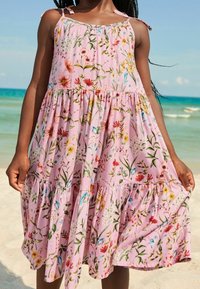 Ein junges Mädchen in einem fließenden pinken Kleid mit bunten Blumenmustern steht am Strand und hält die Seiten ihres Kleides fest.