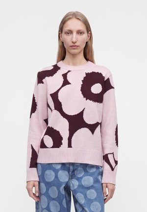 Kvinde iført en lyserød sweater med store mørke blomstermønstre og blå jeans med store lyseblå polkaprikker, stående mod en ensfarvet baggrund.