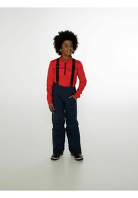 Maglia rossa a maniche lunghe con colletto a zip, abbinata a pantaloni da neve blu navy con bretelle nere regolabili e vestibilità comoda. Scarpe nere con dettagli.