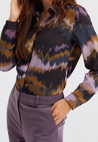 Kvinna som bär en mönstrad skjorta med knappar i brunt, svart och lila, matchat med högmidjade lavendelbyxor, en hand i fickan och guldringar.