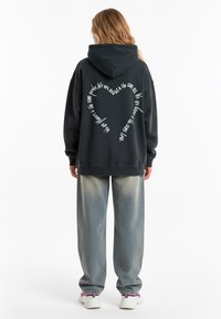 Felpa nera con cappuccio e vestibilità oversize, caratterizzata da testo bianco a forma di cuore sulla schiena. Abbinata a jeans di denim blu chiaro a gamba larga.