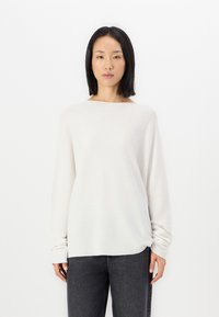 Light beige long-sleeve top in soft, textured material with a boat neckline and gathered detail at the front, paired with dark gray trousers.