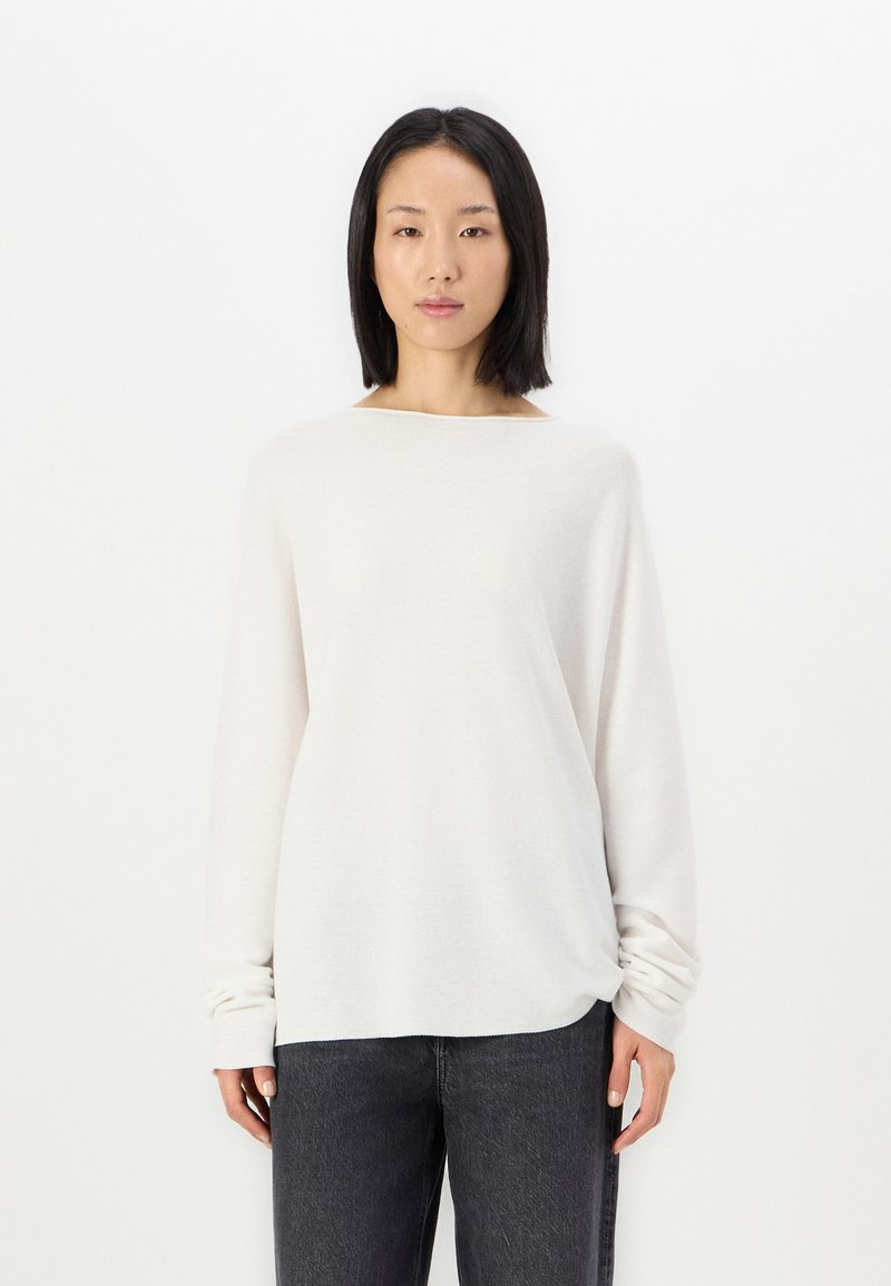 Light beige long-sleeve top in soft, textured material with a boat neckline and gathered detail at the front, paired with dark gray trousers.