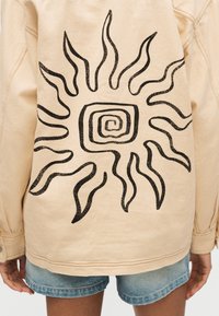 Άτομο που φοράει μπεζ μπουφάν με μαύρο αφηρημένο σχέδιο ήλιου στην πλάτη και ανοιχτά μπλε τζιν σορτς.