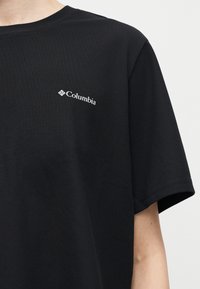 Μαύρο πουκάμισο με κοντά μανίκια και λευκό λογότυπο Columbia στο αριστερό στήθος, που φοριέται από έναν άνθρωπο μπροστά σε απλό ανοιχτό φόντο.