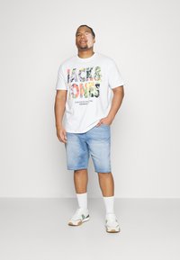 Witte t-shirt met kleurrijke bloemen tekst "JACK & JONES," gecombineerd met lichtblauwe denim shorts en witte sneakers met groene accenten.