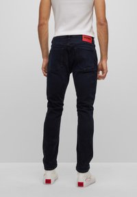 Jeans de mezclilla oscura con dos bolsillos traseros, con una cintura de marca roja. Llevados con zapatillas blancas con acentos rojos.
