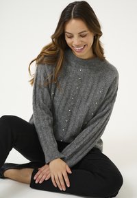 Pull en maille grise avec une texture côtelée, orné d'accents en perles disposés en motif, associé à un pantalon noir.