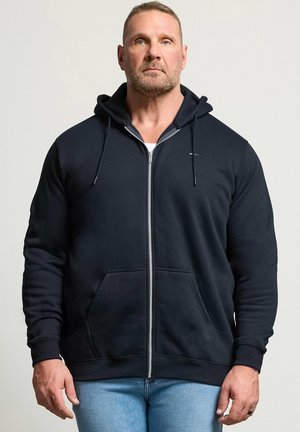 Homme aux cheveux courts et à la barbe debout face à l’avant, portant un sweat noir zippé et un jean bleu clair sur un fond uni.
