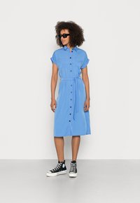 Robe chemise bleu clair à manches courtes, fermeture à boutons sur le devant, deux poches poitrine et taille nouée ; associée à des baskets montantes noires.